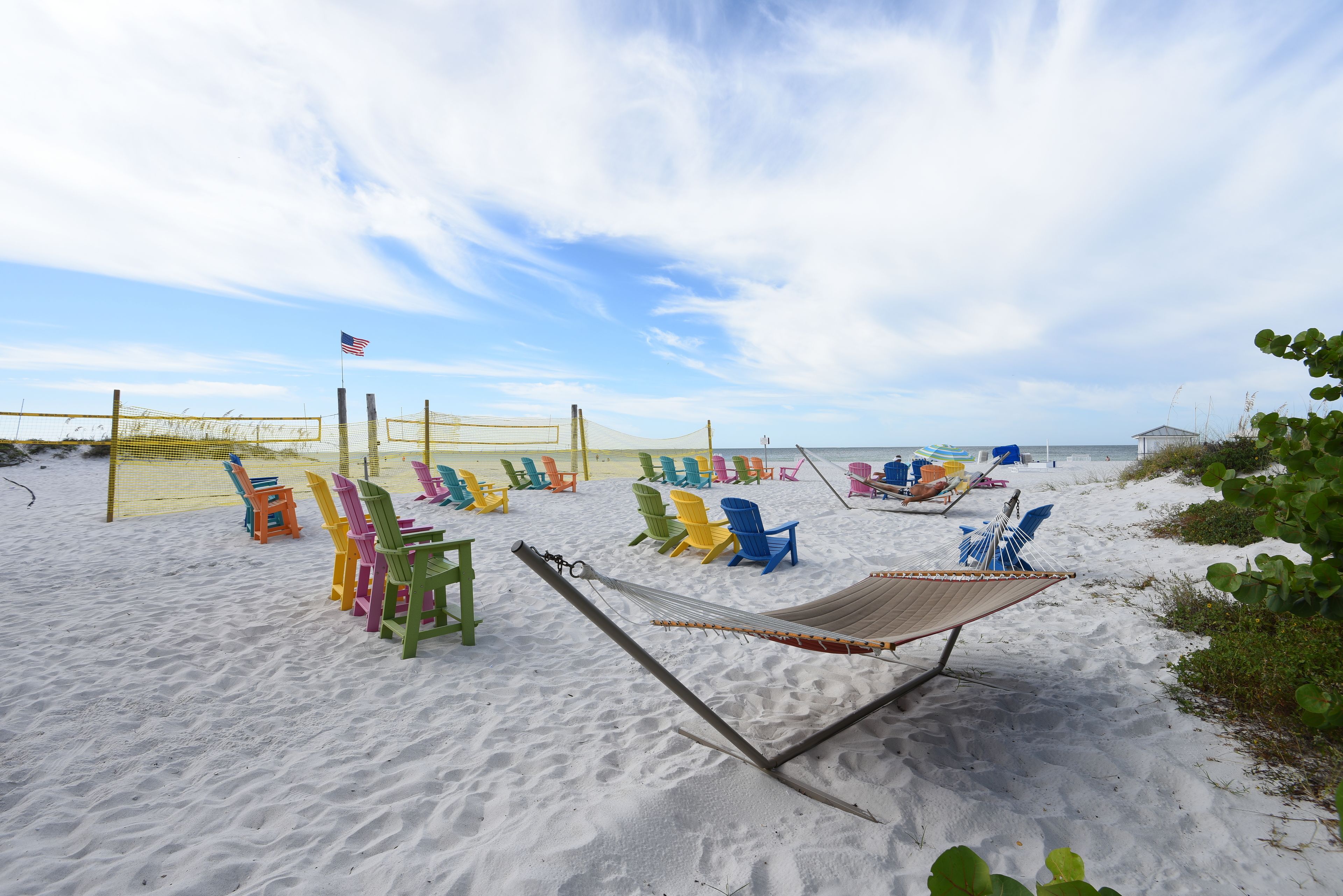Uma praia com cadeiras Adirondack coloridas, uma rede e uma bandeira.