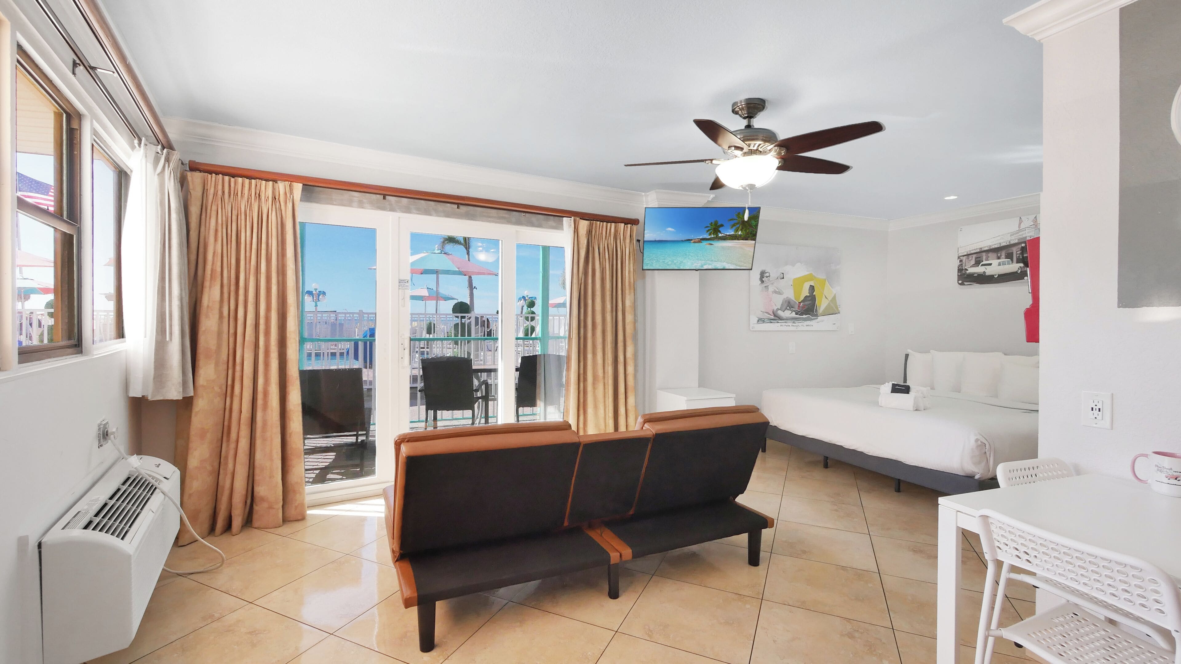 Una camera d'albergo con un letto, un divano, una TV, una ventola a soffitto e un balcone con vista sulla spiaggia.