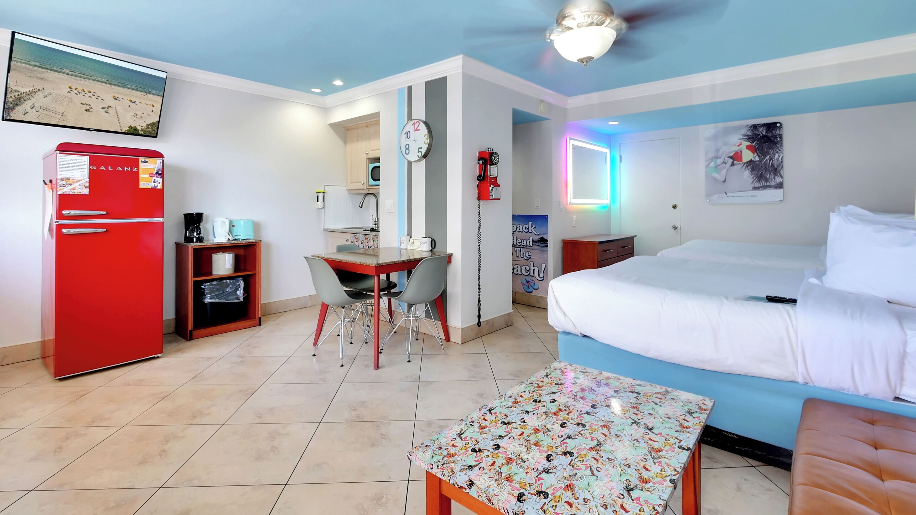 A hotel room with a bed, a desk with a chair, a red refrigerator, a wall clock, and a ceiling fan.
