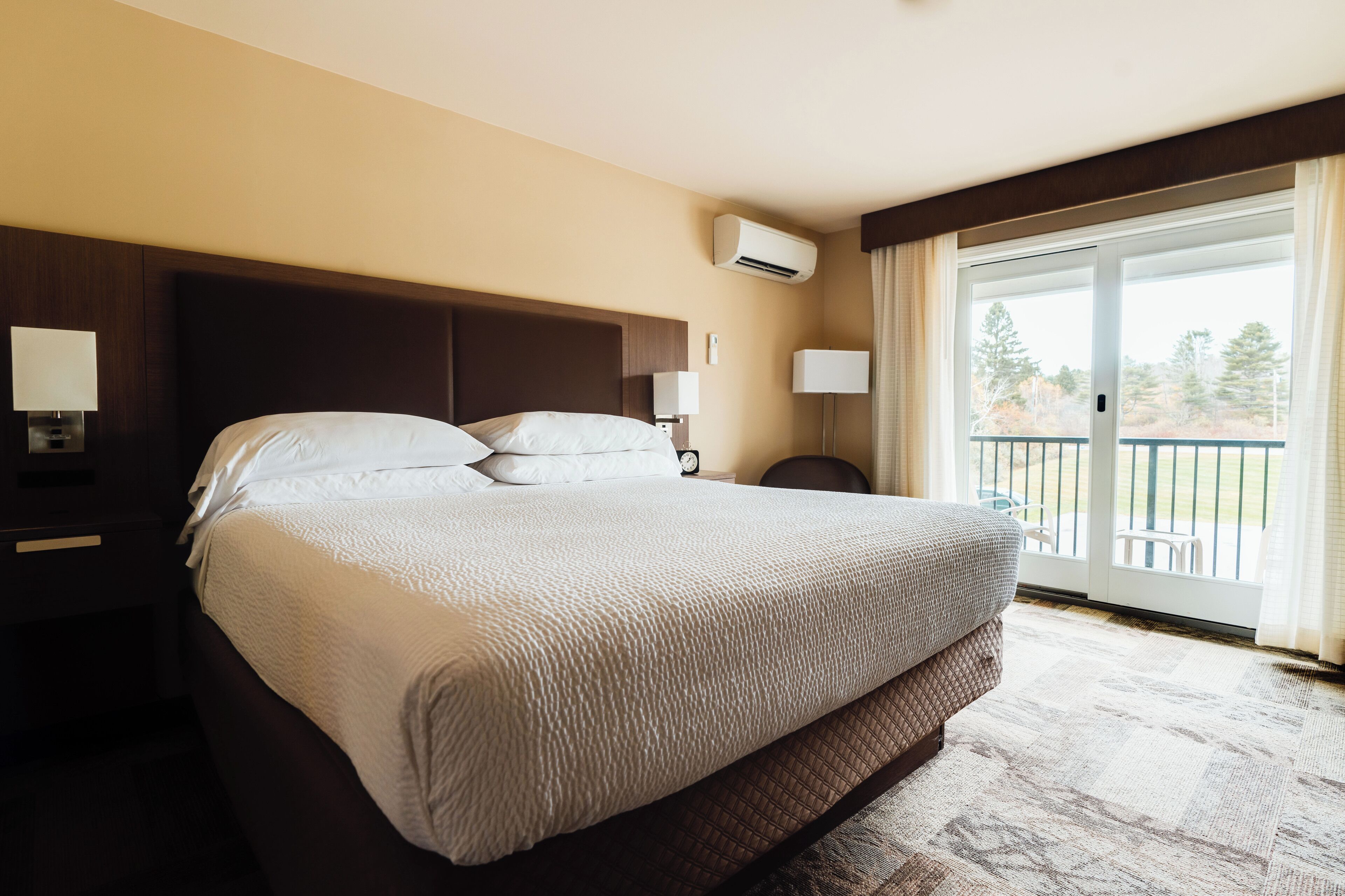 Ein Hotelzimmer mit einem großen Bett, einem Nachttisch mit Lampe, einer Klimaanlage und Blick ins Freie durch eine Schiebetür.