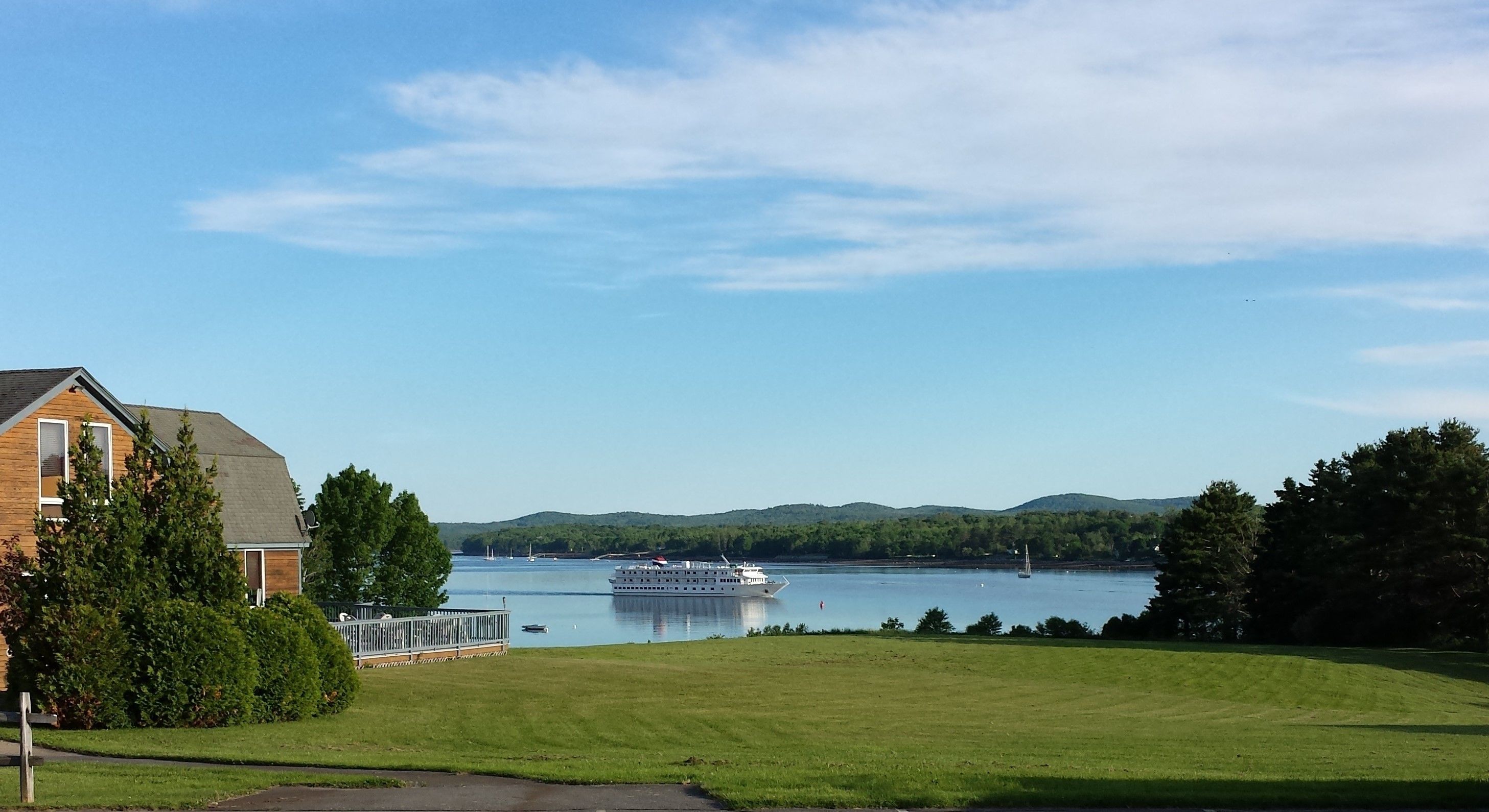 A house with a deck overlooking a lake, a large boat in the water, and a hilly landscape in the distance.