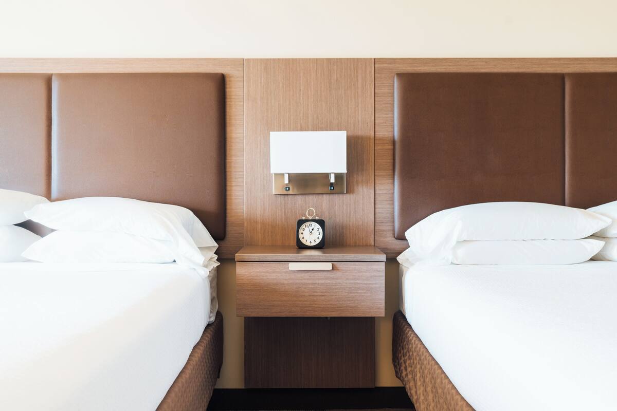 Ein Hotelzimmer mit zwei Betten, einem hölzernen Kopfteil, einem Nachttisch mit einer Uhr und einer Wandlampe.