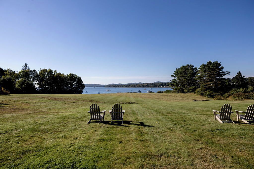 A grassy field with three Adirondack chairs facing a body of water with boats.