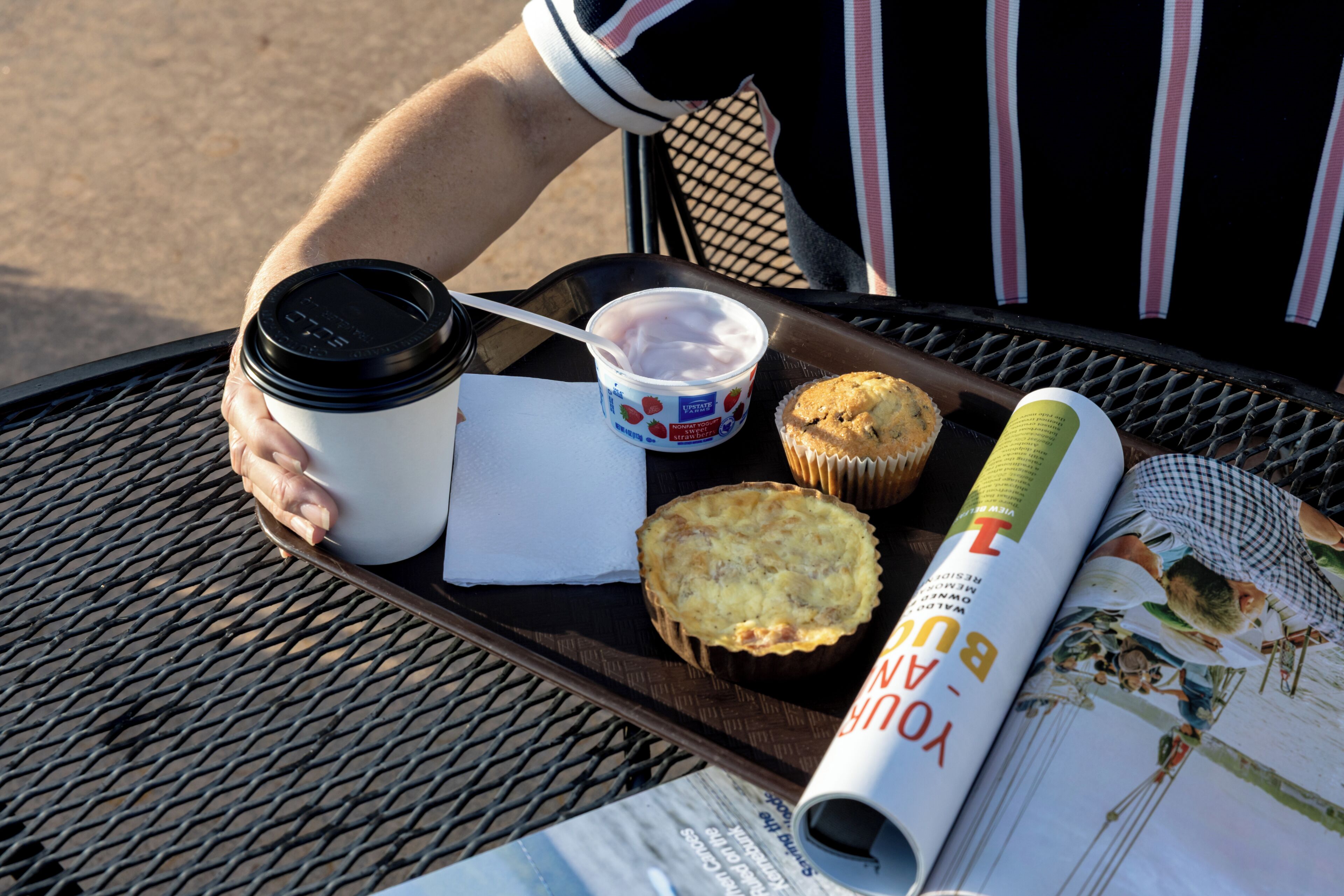 A tray with a coffee cup, muffin, and a rolled-up magazine on a metal table.