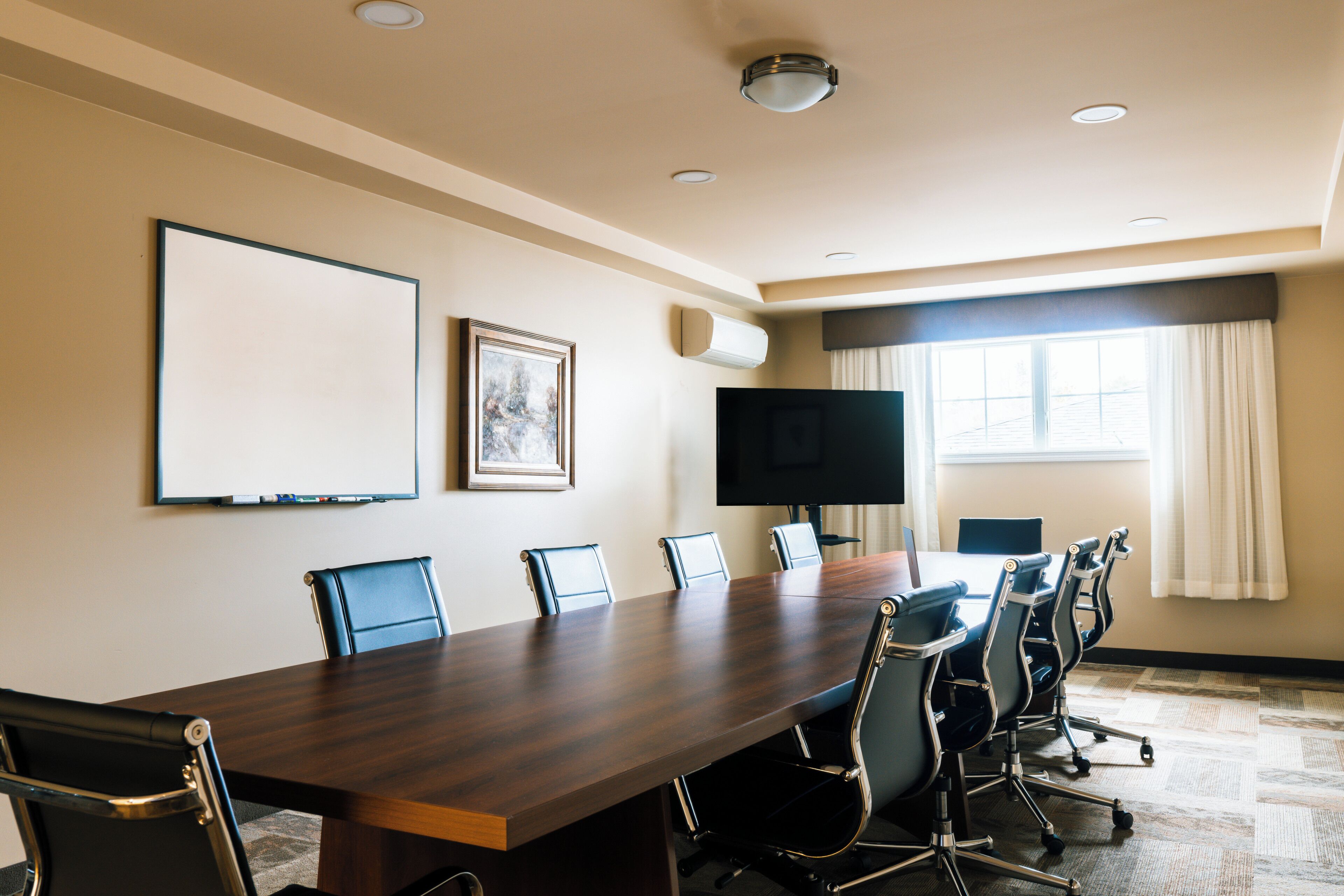 A conference room with a long table, six chairs, a flat-screen TV, a whiteboard, and a window with curtains.