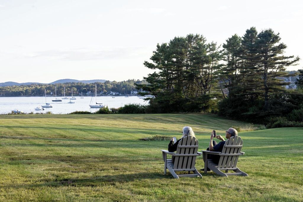 Two people seated on Adirondack chairs facing a lake with boats, surrounded by trees and a grassy area.