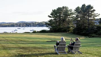 Two people seated on Adirondack chairs facing a lake with boats, surrounded by trees and a grassy area.