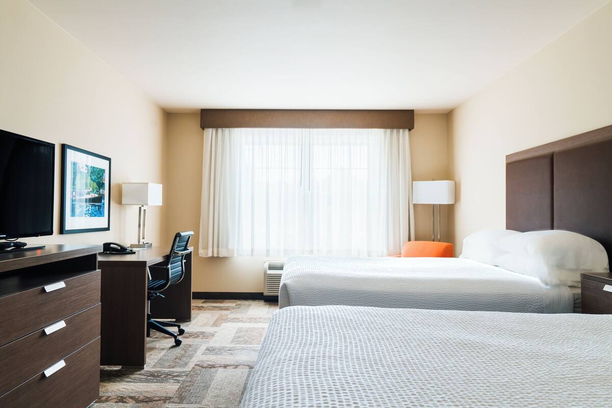Ein Hotelzimmer mit zwei Betten, einem Schreibtisch, einem Stuhl, einem Fernseher und einem Fenster mit Vorhängen.