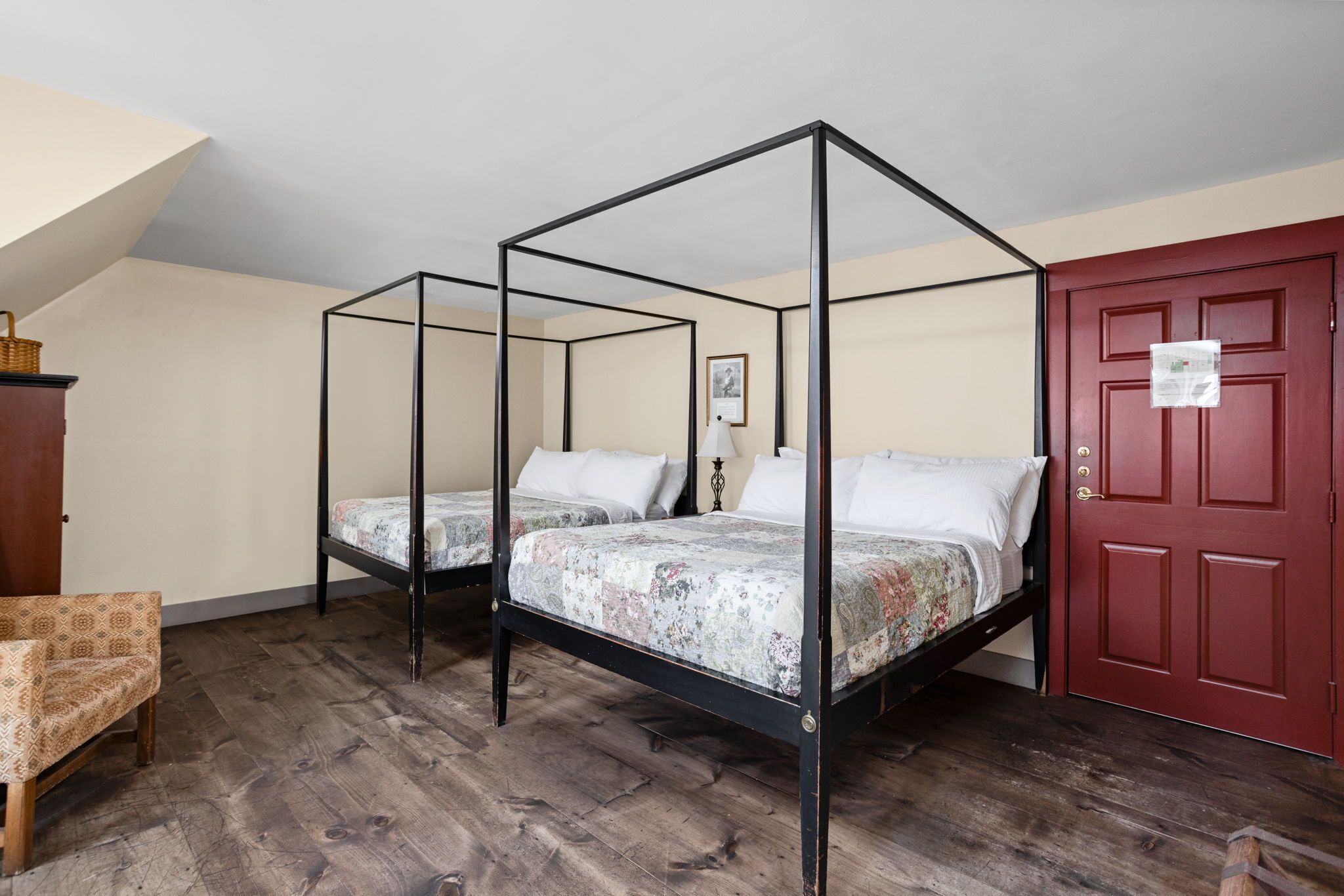 A room with two four-poster beds, a wooden floor, a red door, and a small chair.