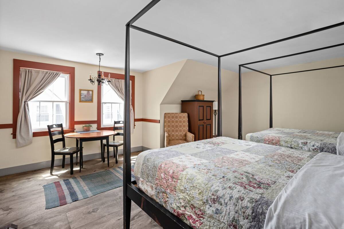 A four-poster bed with a floral quilt, a wooden desk with chairs, and a window with curtains.
