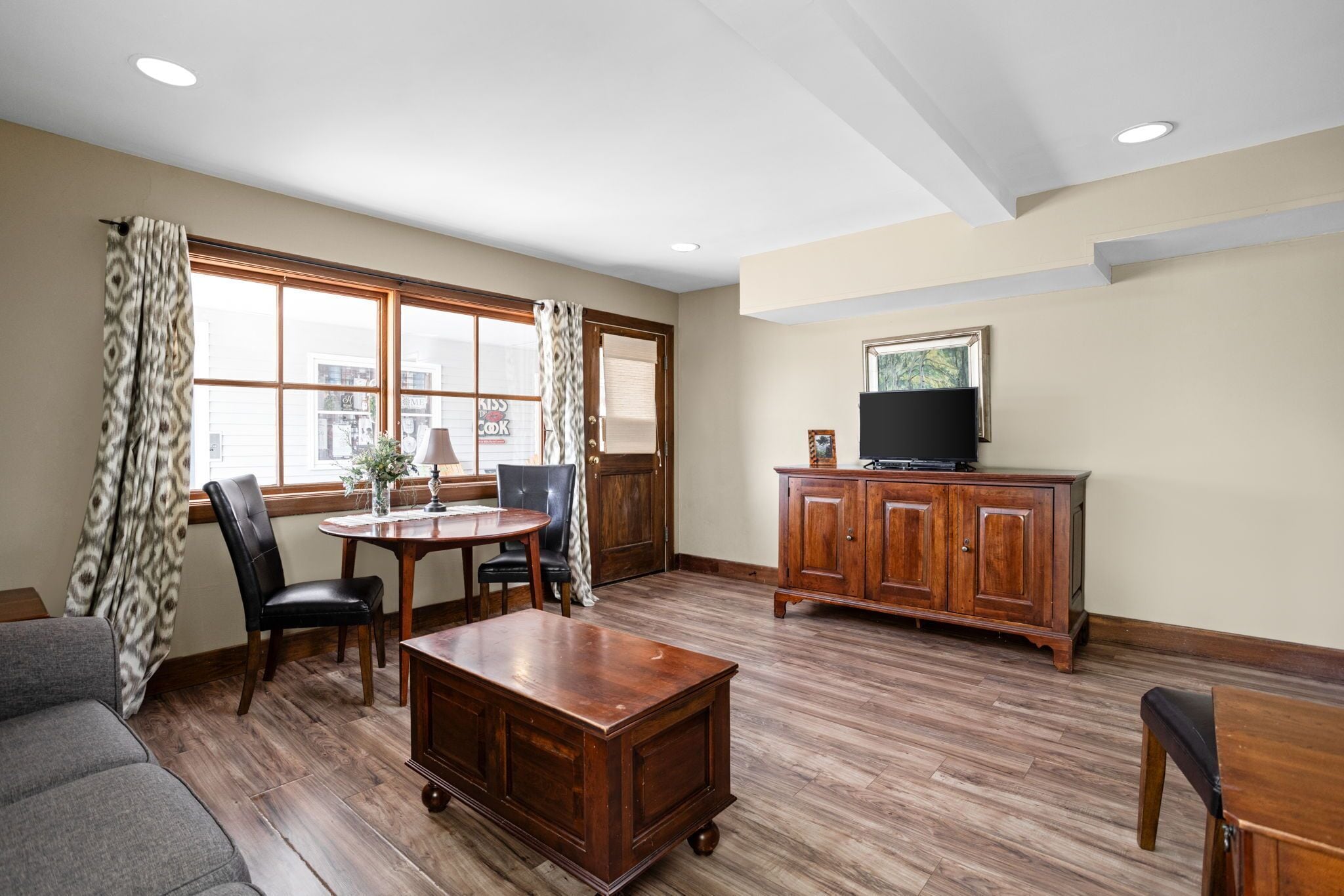 A spacious living room with a wooden floor, a dining table with chairs, a sofa, a cabinet, and a television on a stand.