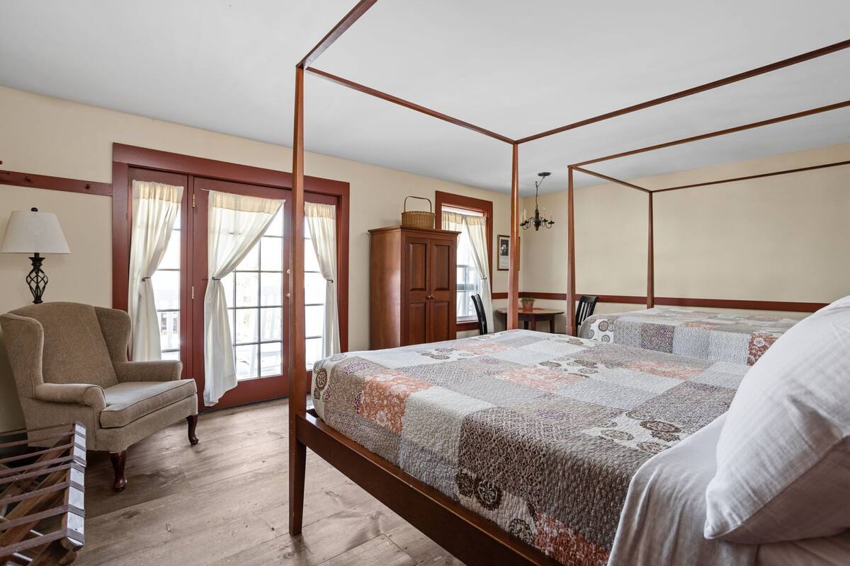 A four-poster bed with a patterned bedspread, a wooden chair, and a desk with a lamp.