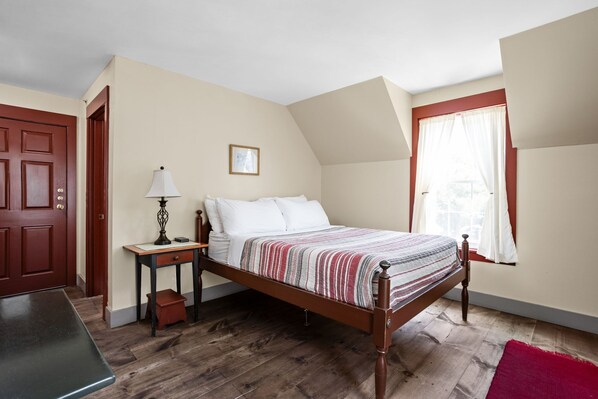 A bedroom with a wooden bed, a nightstand, a desk, a chair, a red rug, a door, and a window with curtains.