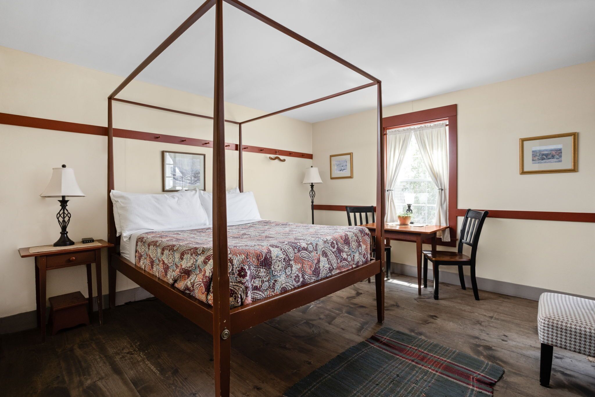 A four-poster bed with a patterned bedspread, a wooden nightstand with a lamp, a chair, a small table with a lamp, and a window with curtains.