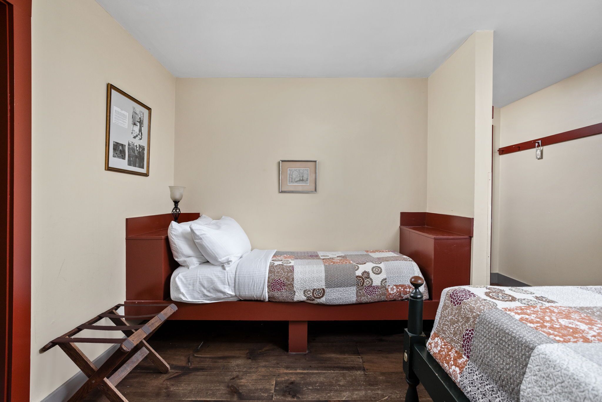 Una stanza piccola con un letto singolo, una testiera e una panca di legno, e un tavolino. Due quadri sono appesi alla parete.