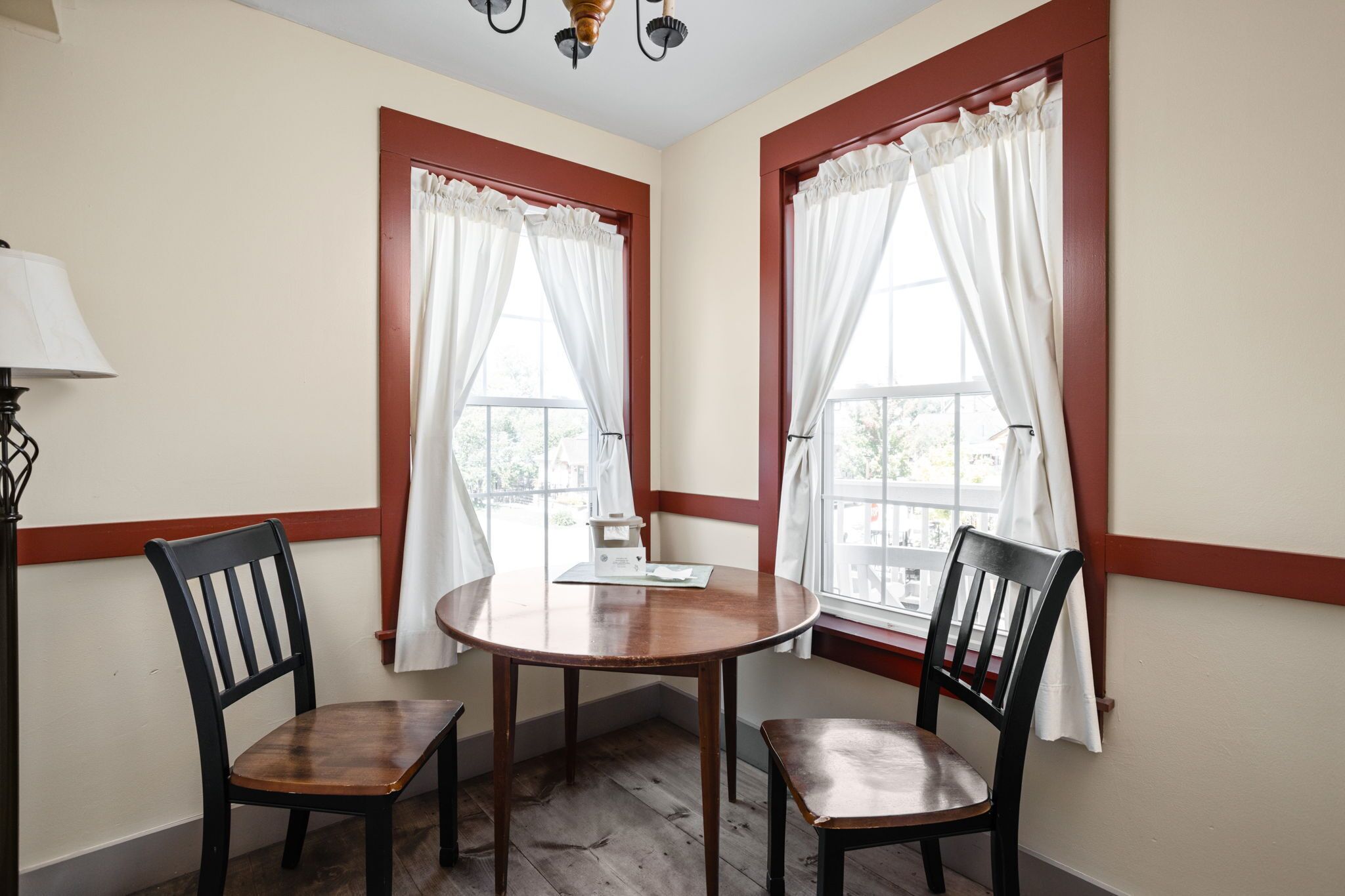 A room with a round wooden table and two chairs, two windows with white curtains, and a lamp on the left.