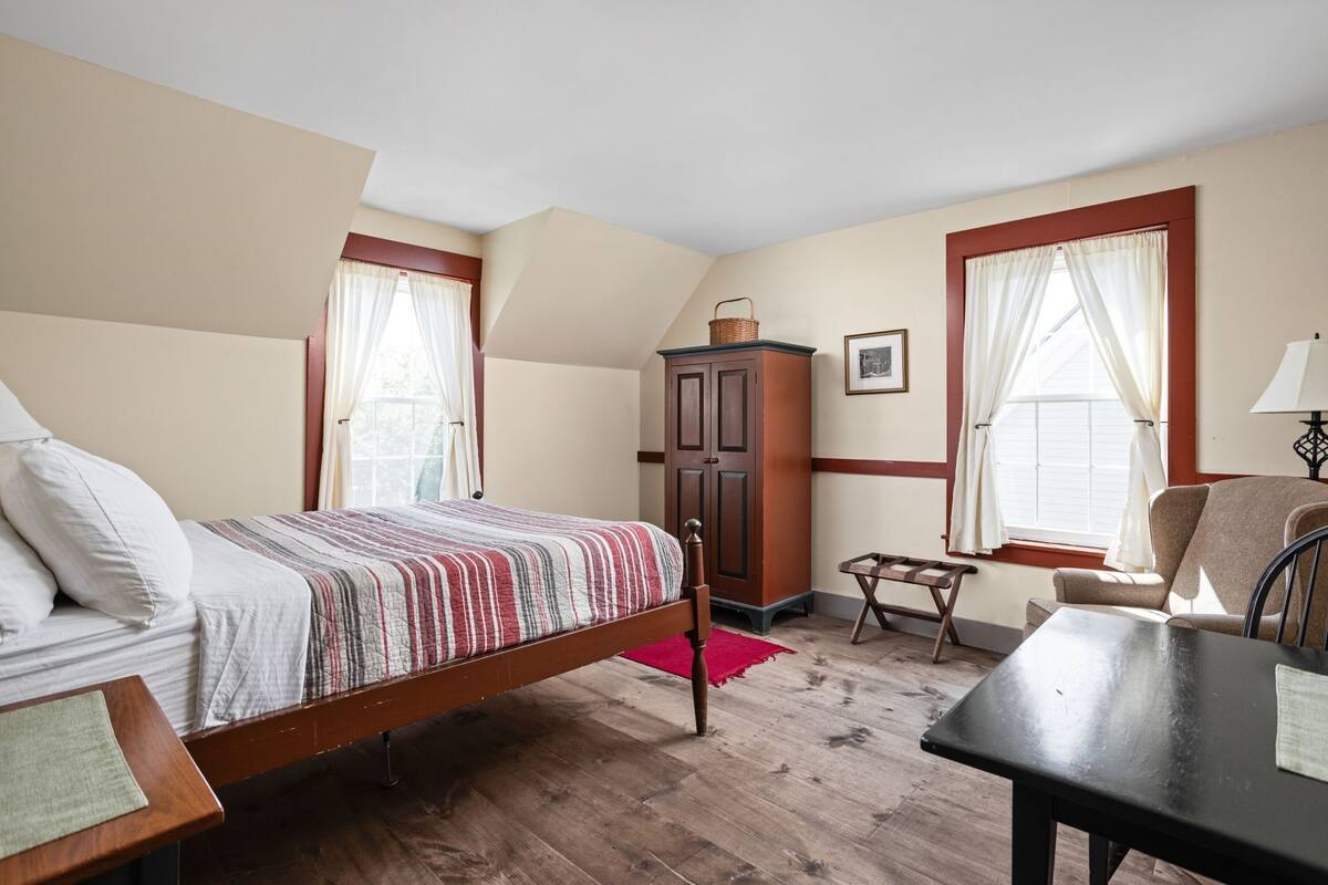 A bedroom with a wooden bed, a small table, a chair, a cabinet, and a window with curtains.