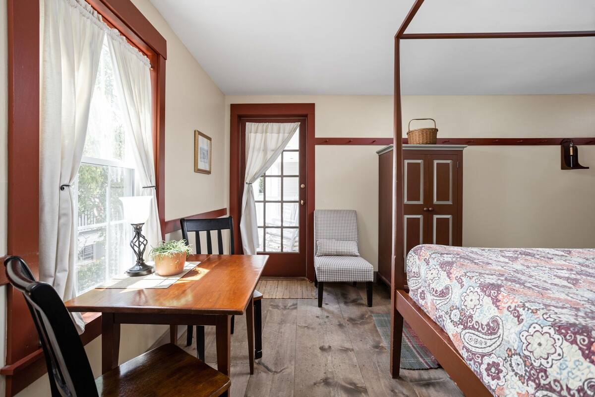 A bedroom with a wooden four-poster bed, a checkered chair, a wooden table with a potted plant, and a window with curtains.