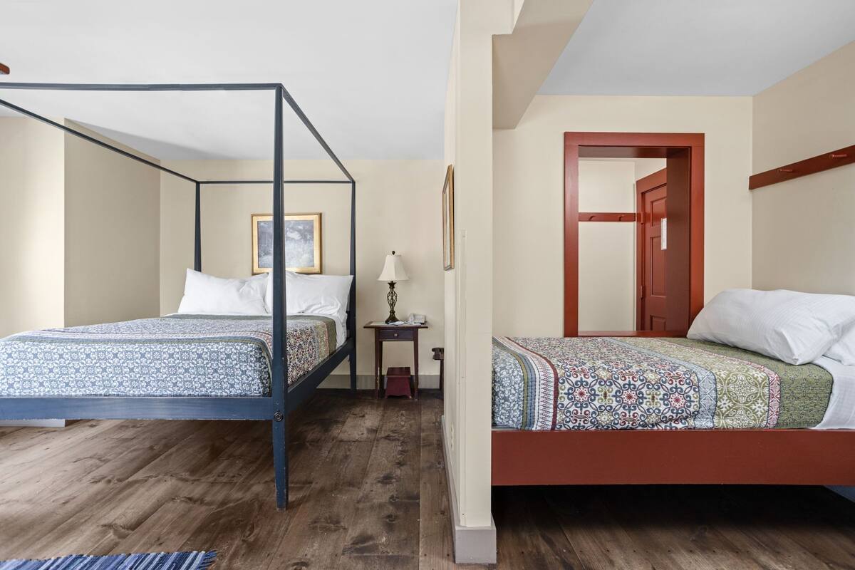 A room with two beds, a wooden floor, a small table with a lamp, and a door leading to another room.
