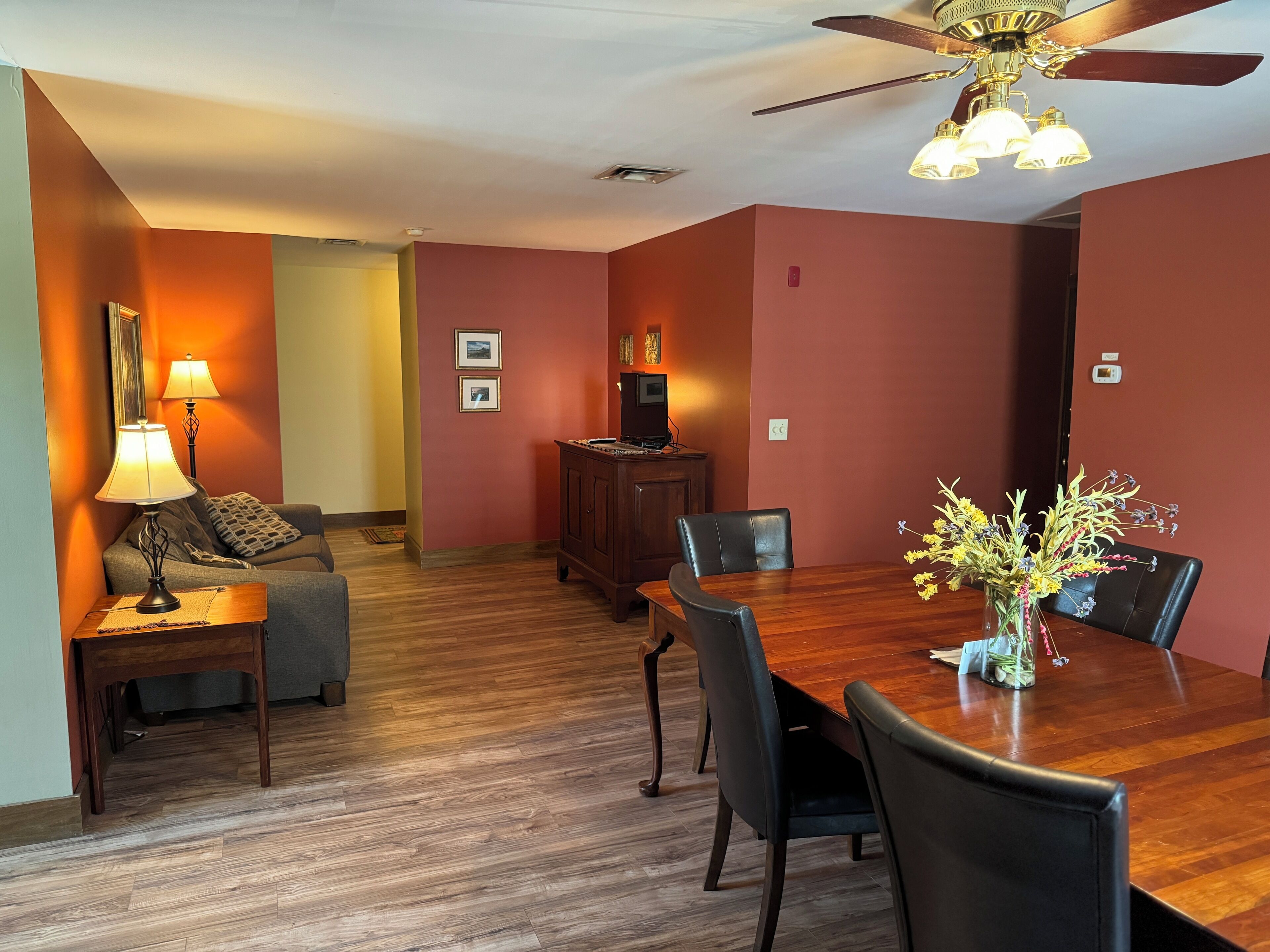 A dining area with a wooden table, black chairs, a vase with flowers, a ceiling fan, and a lamp on a side table.