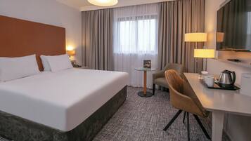 Ein Hotelzimmer mit einem großen Bett, einem Schreibtisch mit Stuhl, einer Kaffeemaschine und einem Fernseher.