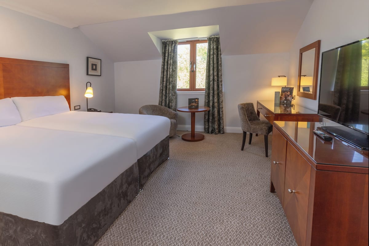 Een hotelkamer met een groot bed, een bureau met een televisie, een klein tafeltje met een lamp en een raam met gordijnen.