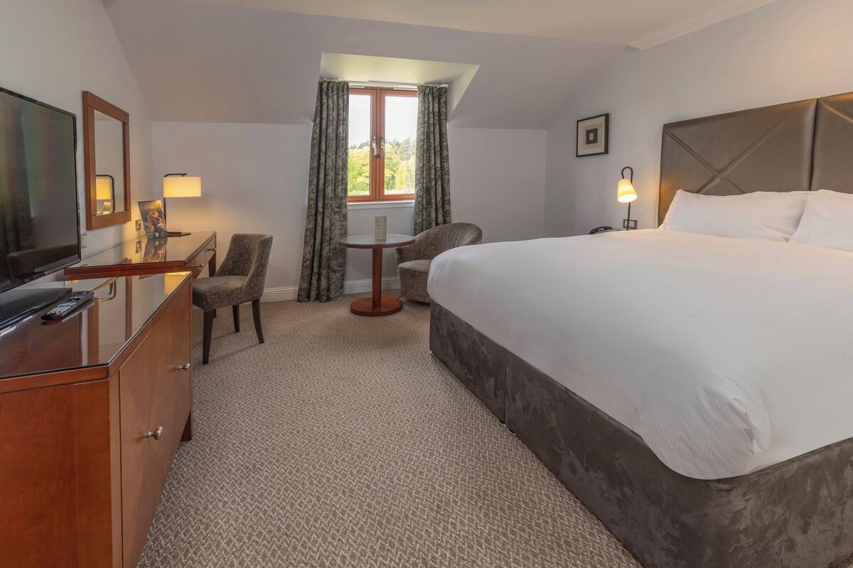 Een hotelkamer met een groot bed, een bureau met een televisie, een stoel, een klein tafeltje en een raam met gordijnen.