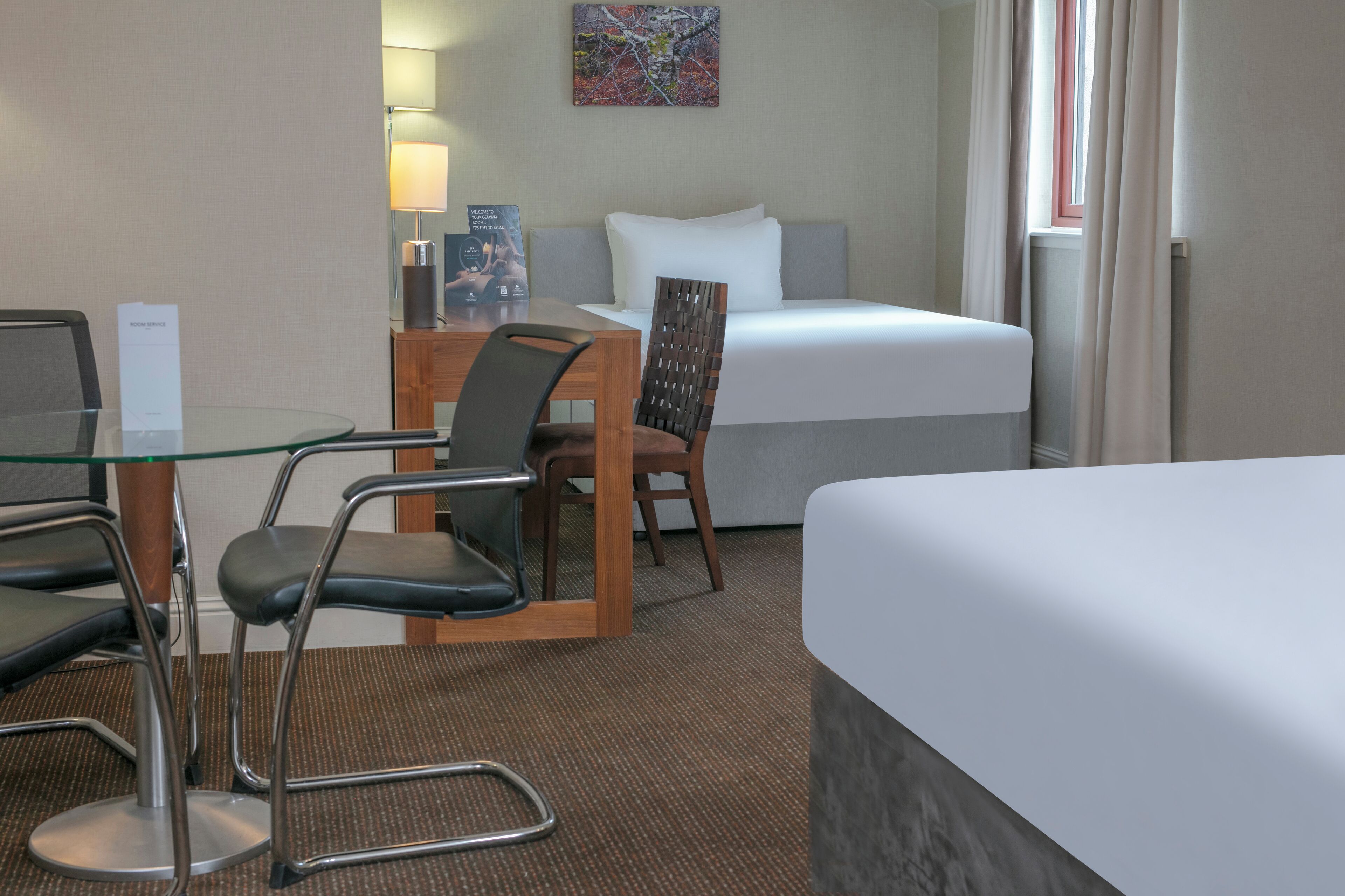 A hotel room with a bed, a glass table, two chairs, a lamp, and a painting on the wall.