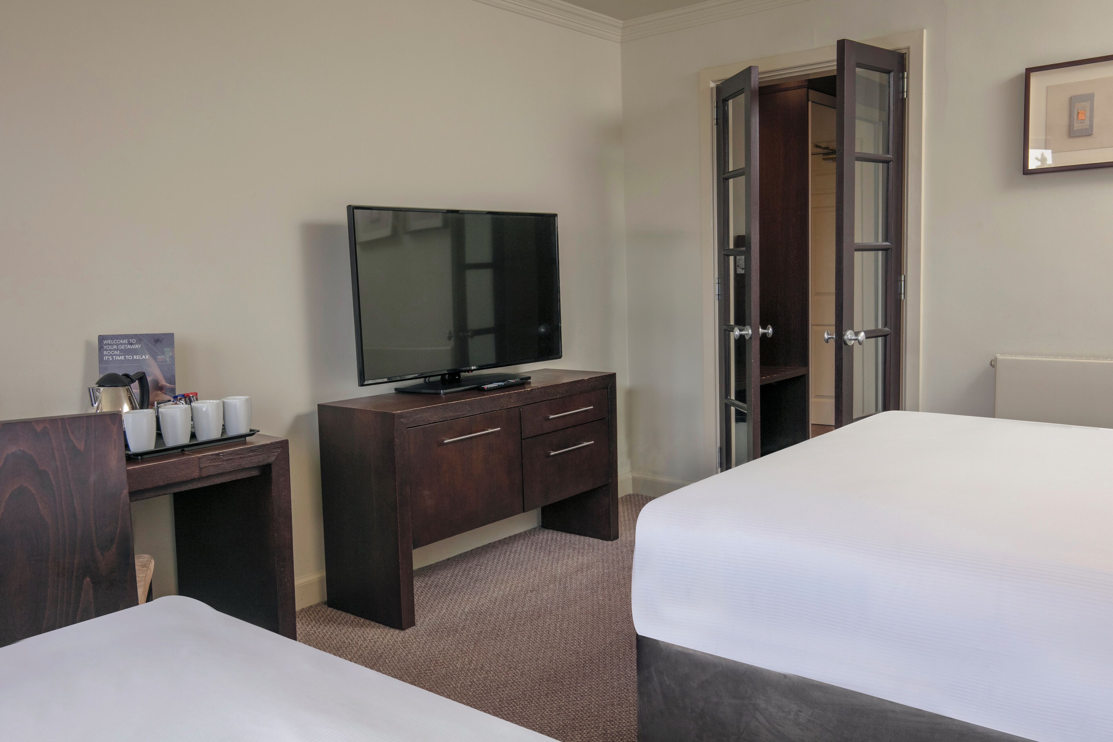 Una habitación de hotel con una cama, un televisor sobre un aparador de madera, un escritorio con cafetera y tazas, y un armario.