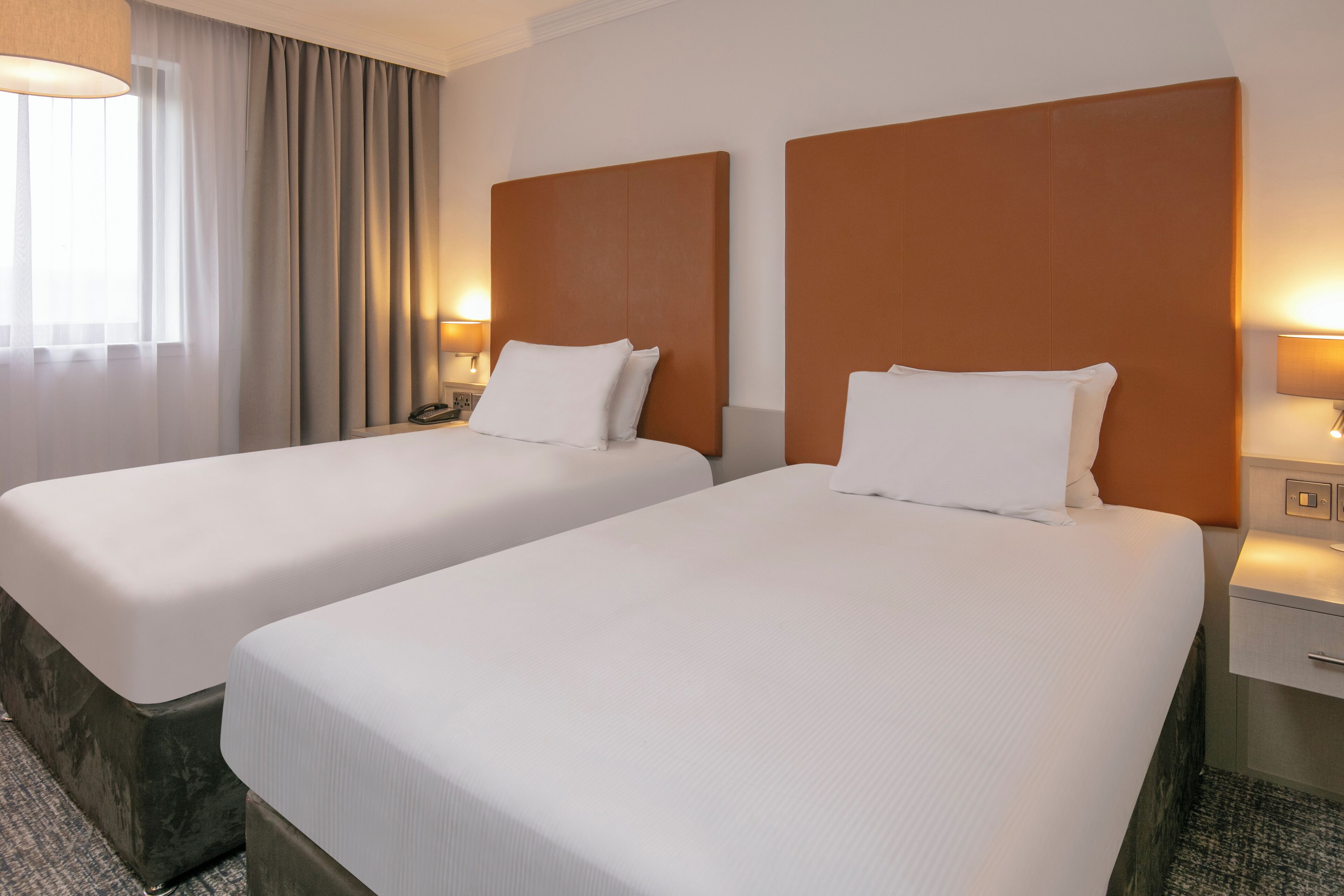 Habitación de hotel con dos camas, cada una con cabecero de madera y ropa de cama blanca. Hay mesitas de noche con lámparas y un teléfono.