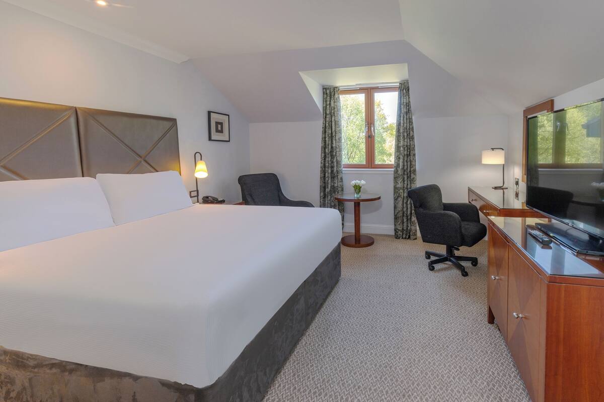 Een hotelkamer met een groot bed, een bureau met televisie, een stoel, een klein tafeltje met een vaas en een raam met gordijnen.
