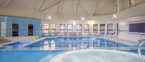 Una piscina interior con varias pistas de natación, una zona de estar con sillas y un techo alto con vigas a la vista.