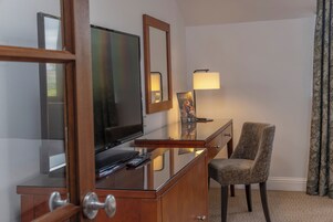 A hotel room with a wooden desk, a television, a chair, a lamp, and a mirror.