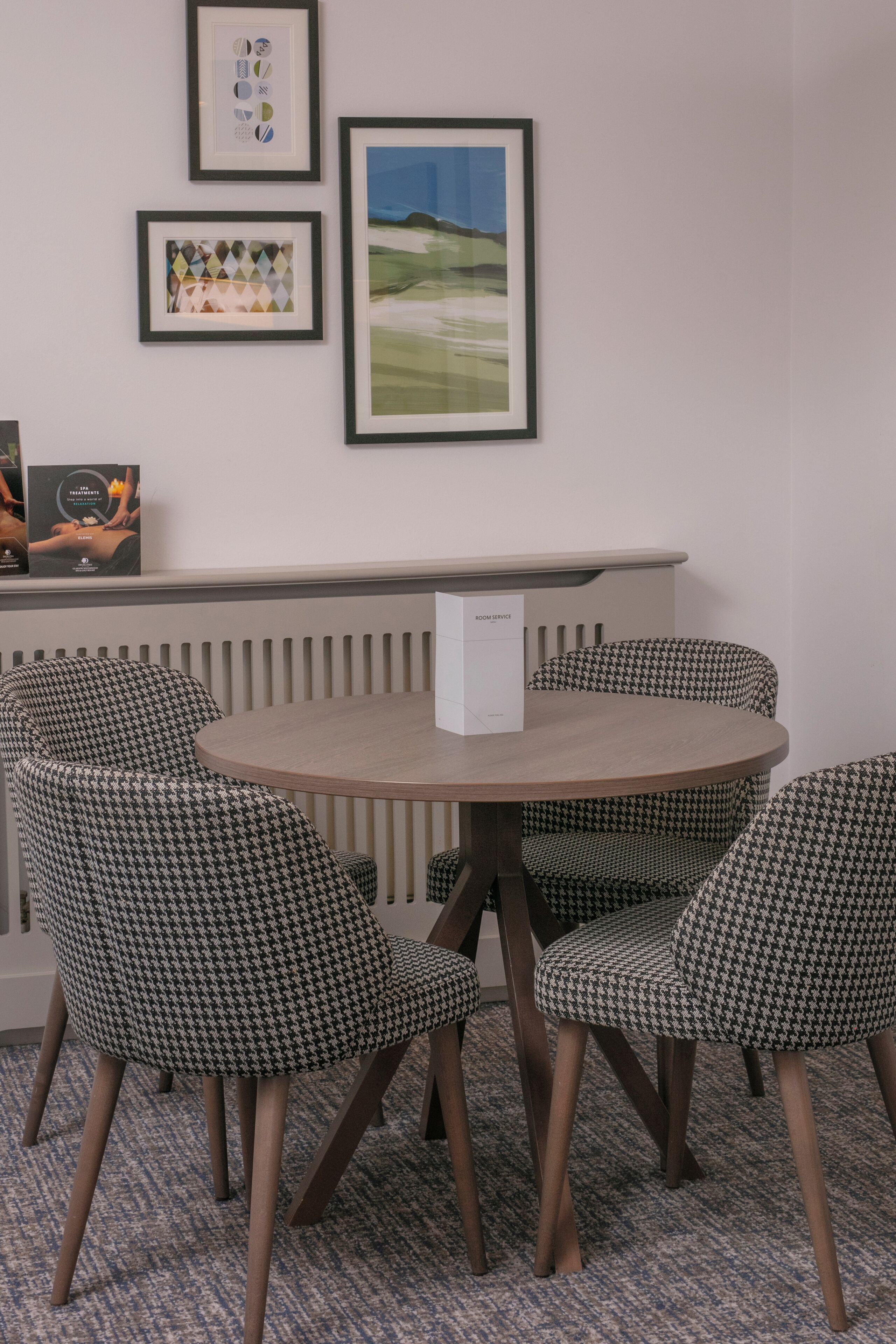 A round table with houndstooth patterned chairs, a framed picture on the wall, and a radiator below.