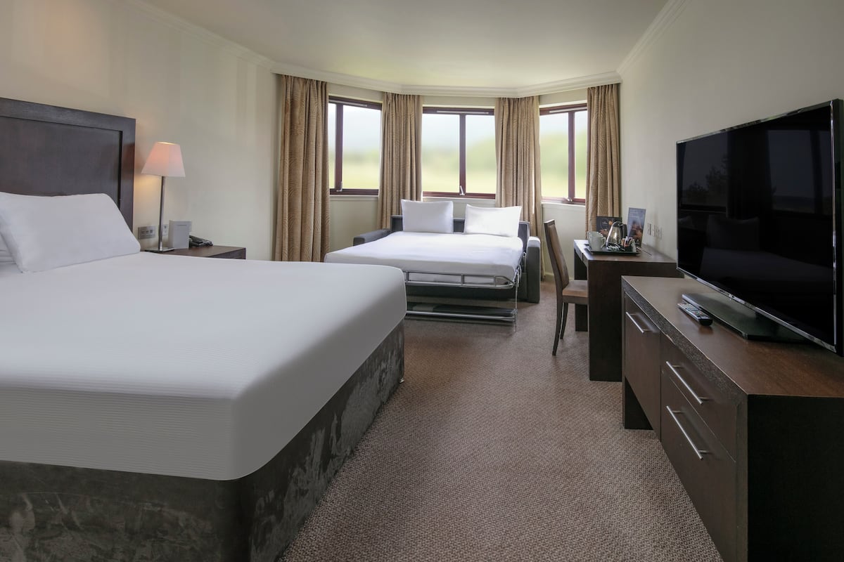 Hotelkamer met een groot bed, een kleiner bed, een bureau met televisie en een raam met gordijnen.