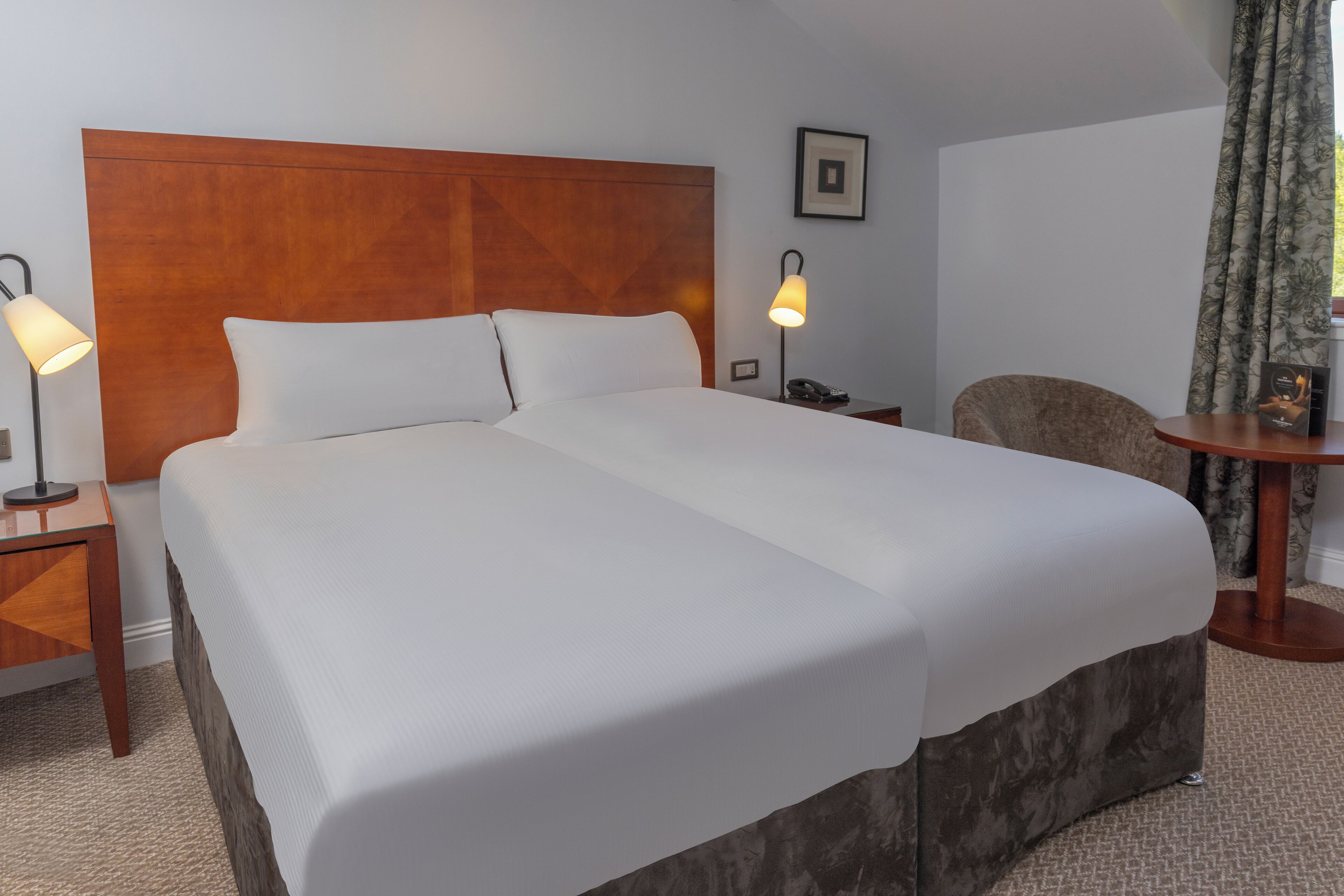 Ein Hotelzimmer mit einem großen Bett, zwei Nachttischlampen, einem Holz-Kopfteil, einem gerahmten Bild an der Wand, einem Sessel und einem kleinen Tisch mit Telefon und Buch.