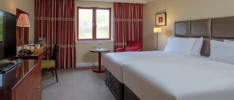 Ein Hotelzimmer mit einem großen Bett, einem Schreibtisch mit Fernseher, einem Sessel, einem kleinen Tisch und Vorhängen.
