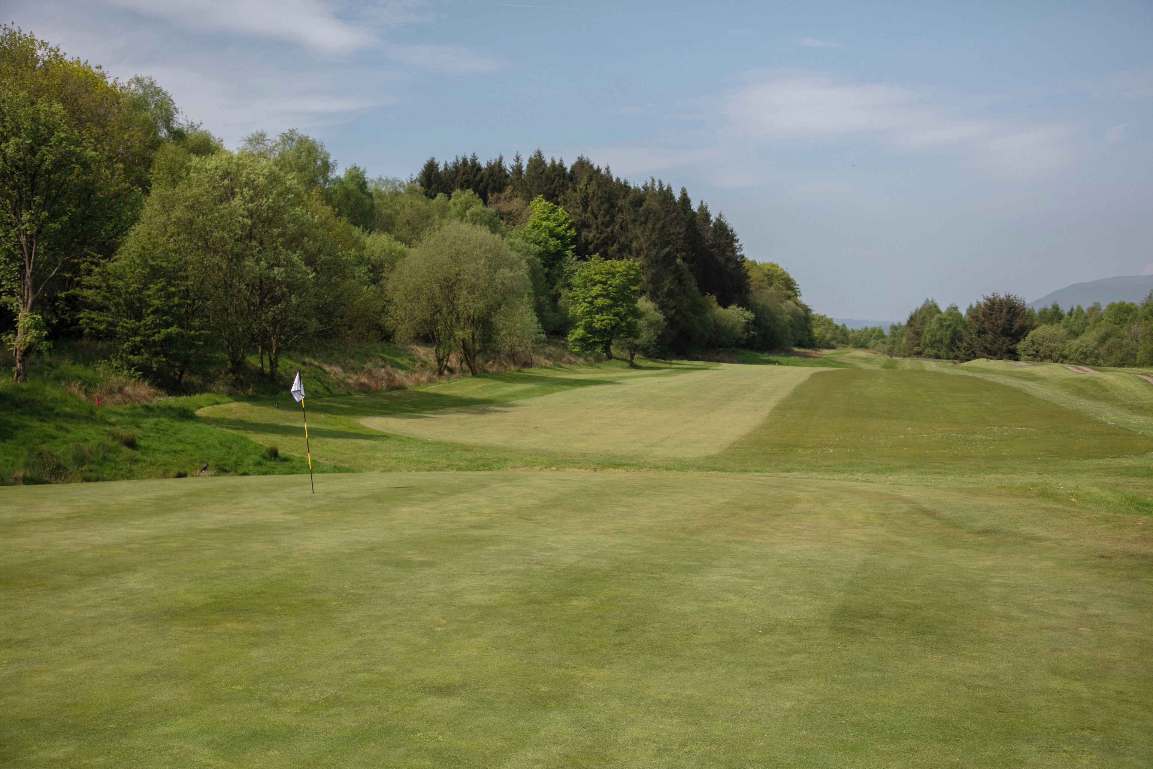 A golf course with a flagstick, surrounded by trees and hills.