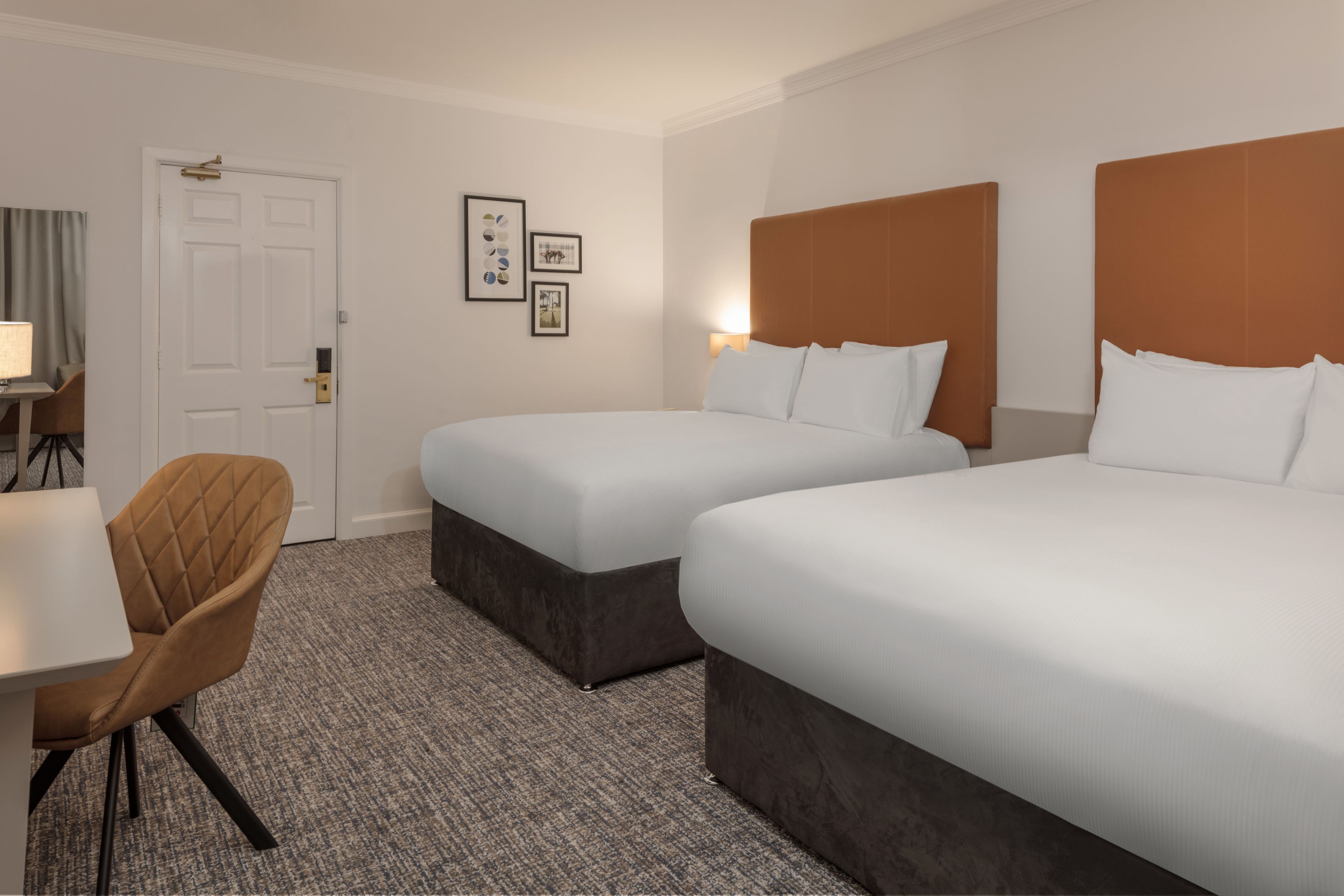 A hotel room with two beds, a desk, a chair, and a door.