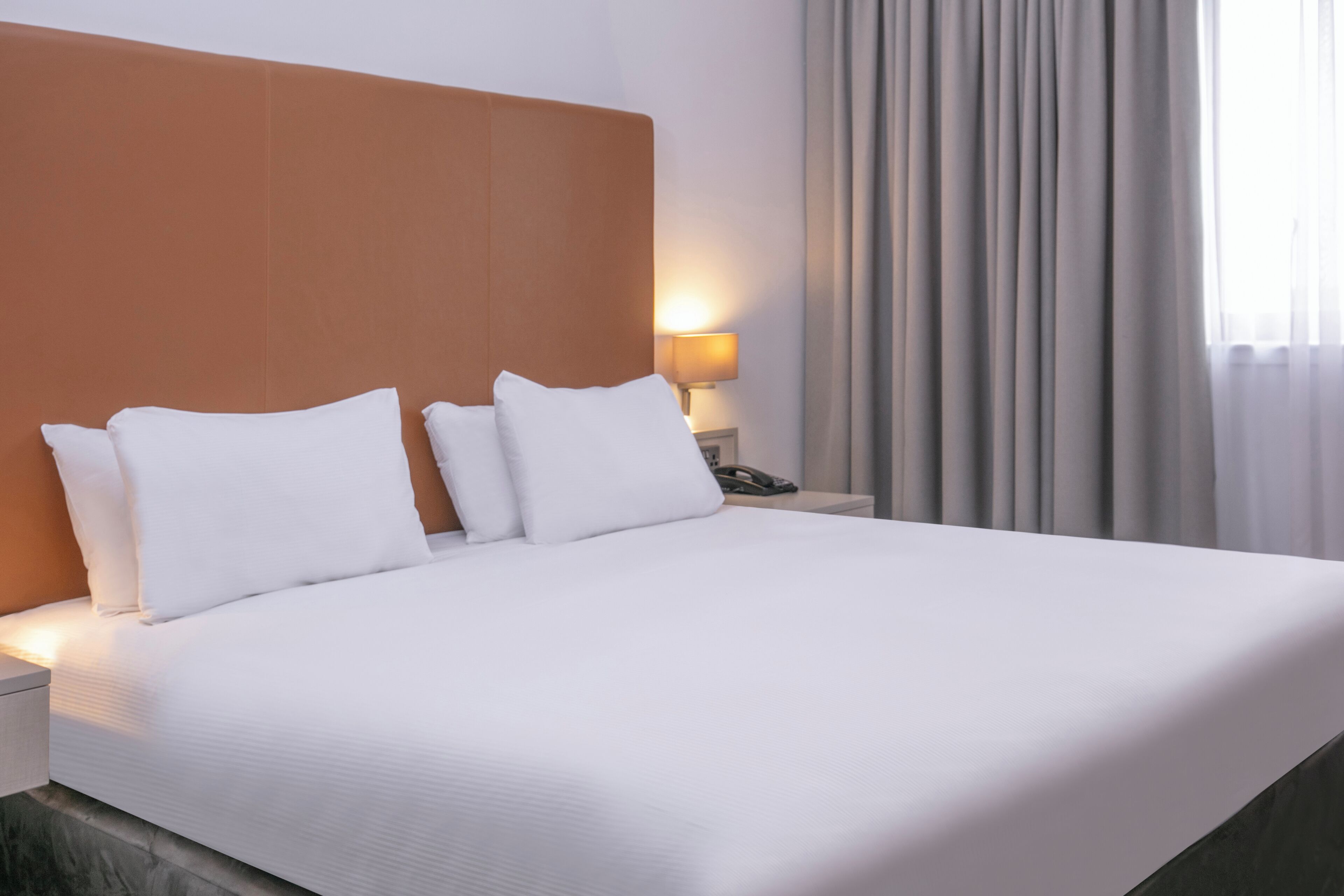 A neatly made bed with white linens and pillows, a wooden headboard, a bedside lamp, and a window with curtains.