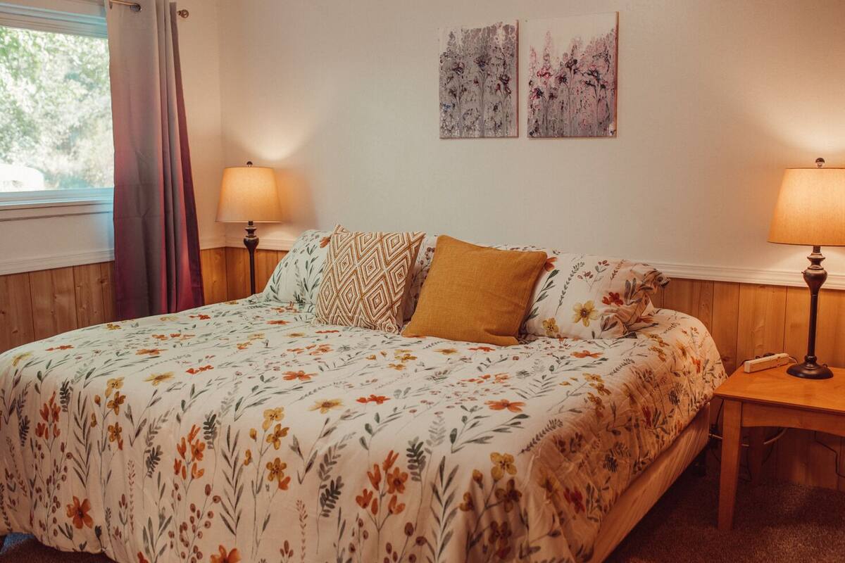 A bedroom with a floral bedspread, two lamps on either side of the bed, and two wall-mounted paintings.