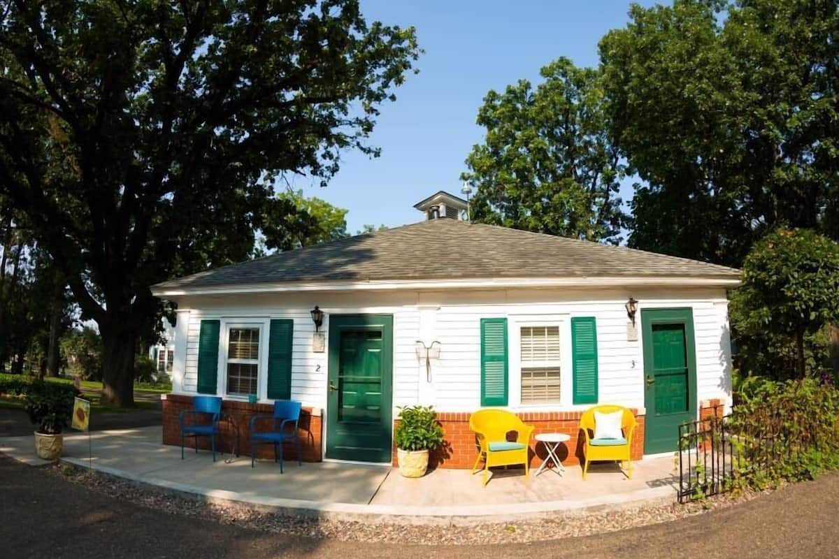 A small, single-story building with green shutters and a white exterior. It features a small porch with yellow chairs and a table, surrounded by potted plants.
