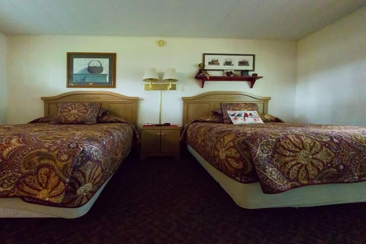 A hotel room with two beds, a nightstand, a lamp, and a shelf with framed pictures.
