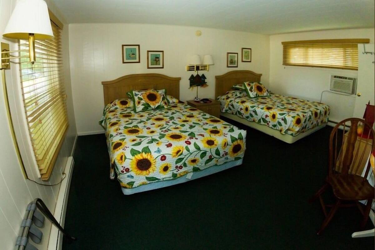 A room with two beds, each with a sunflower-patterned bedspread, a wooden headboard, a nightstand, and a chair.