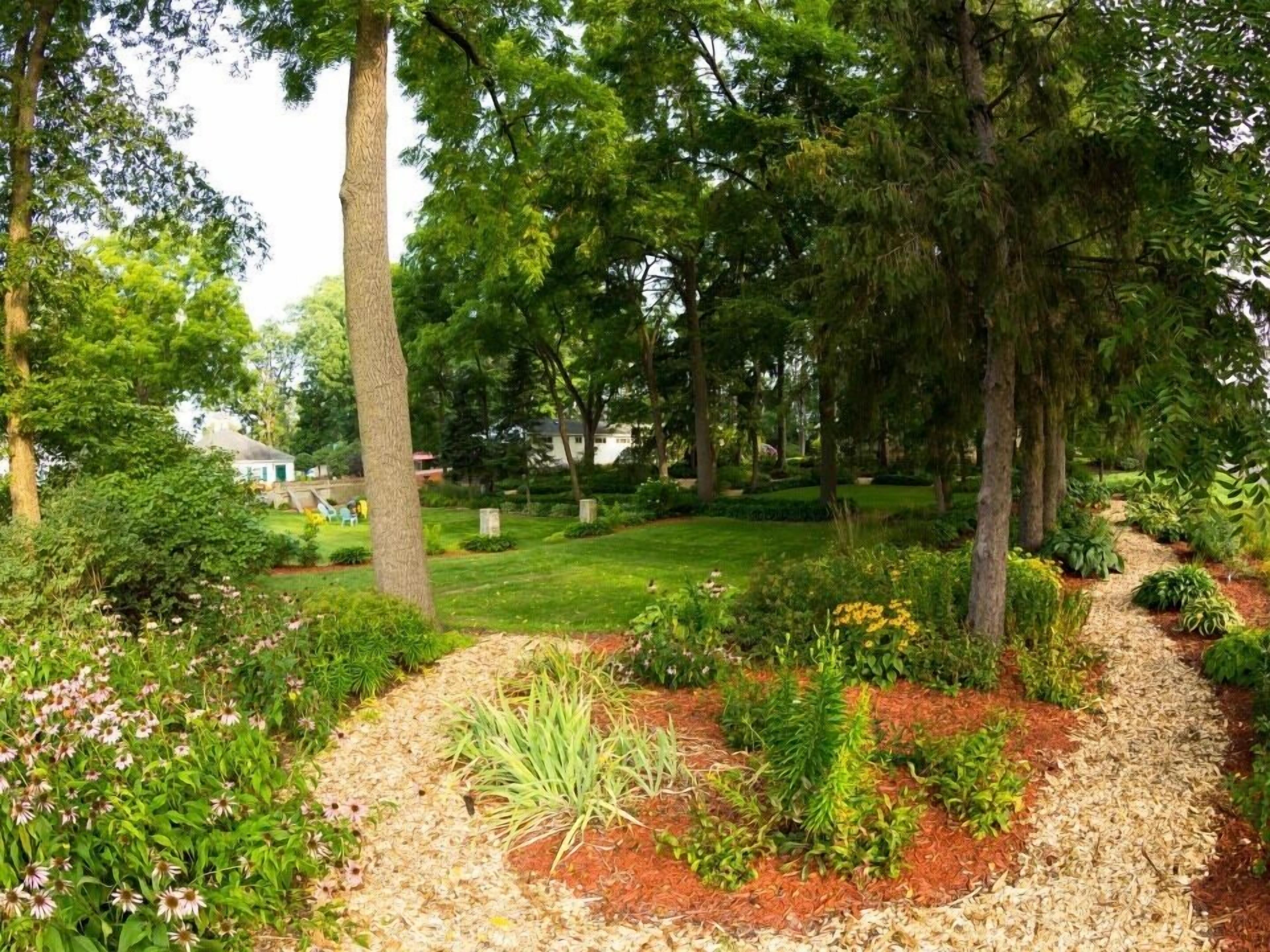 A landscaped garden with a variety of plants and trees, a clear path, and a small building in the background.
