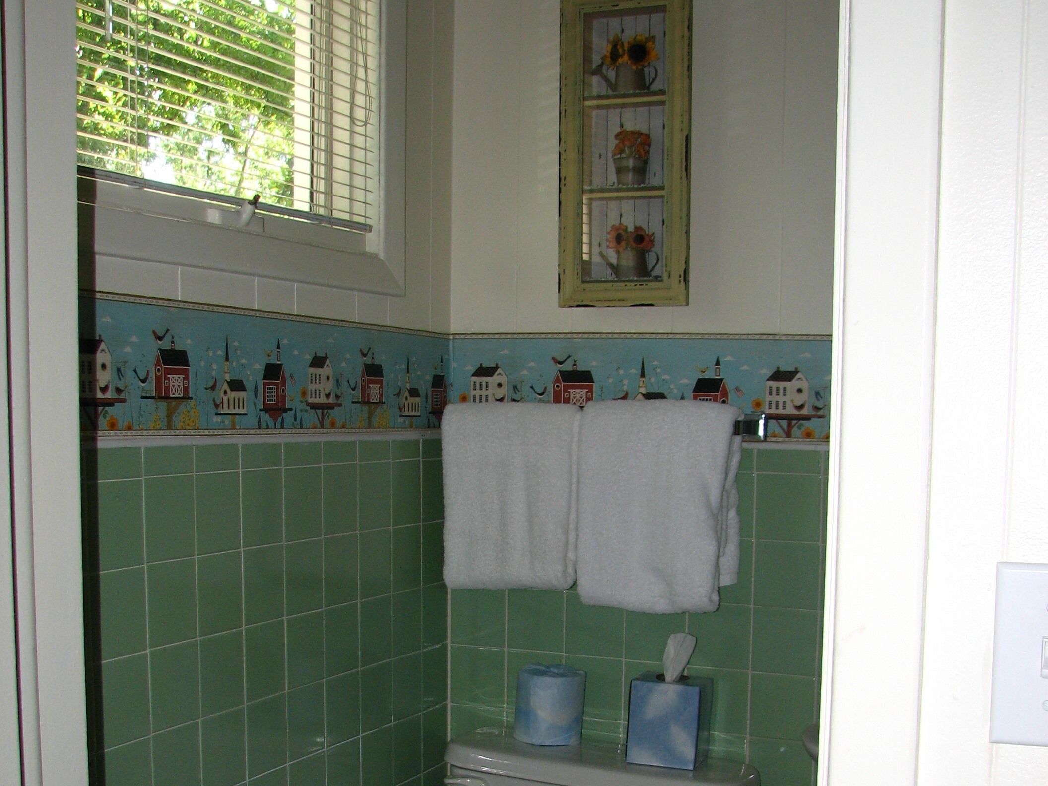 A bathroom with green tiles, a window with blinds, a towel rack with two towels, and a small shelf with a tissue box and toilet paper.