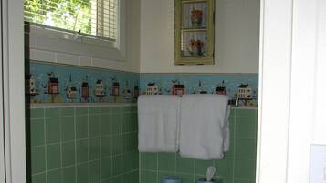 A bathroom with green tiles, a window with blinds, a towel rack with two towels, and a small shelf with a tissue box and toilet paper.