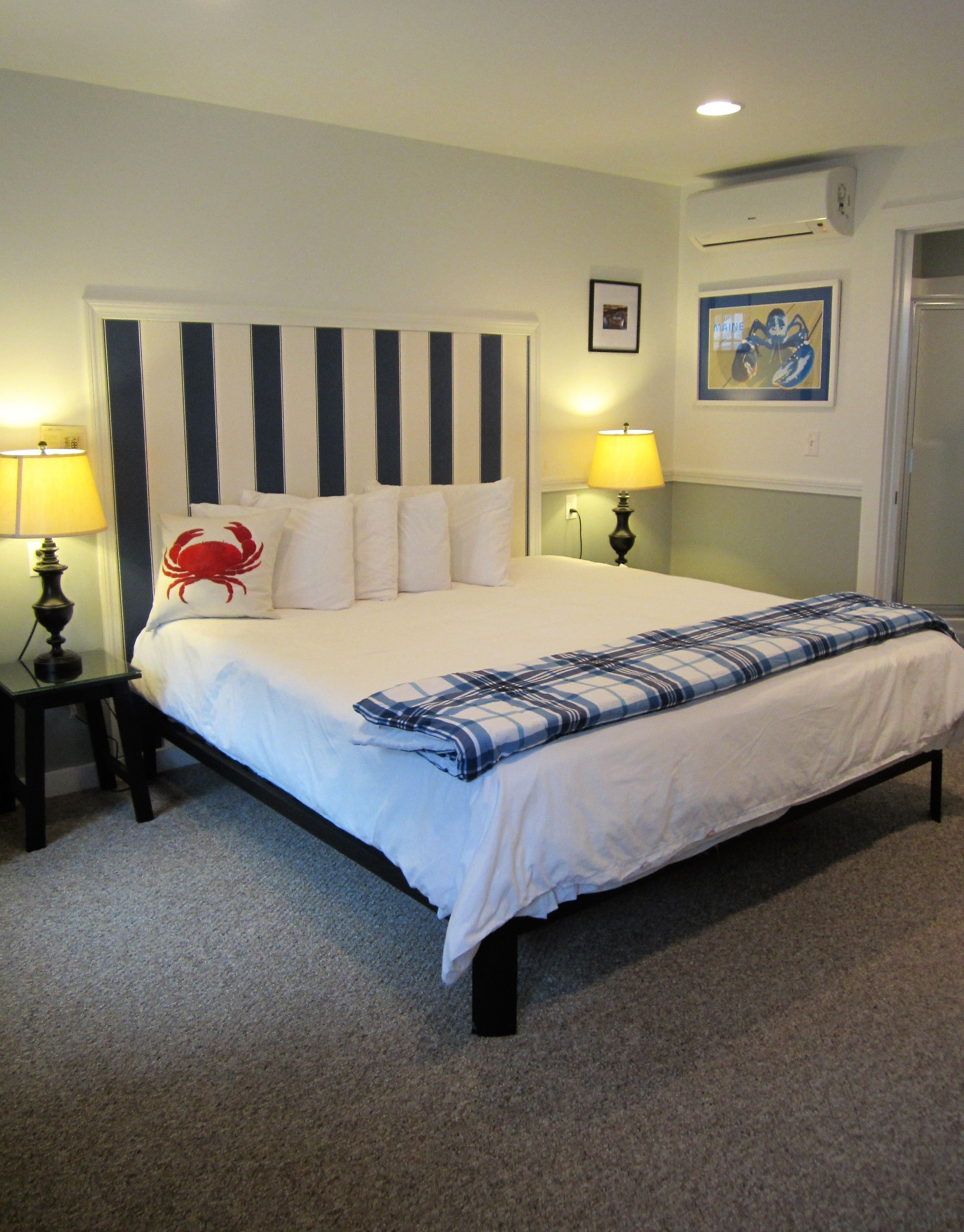 A neatly made bed with a striped headboard, two bedside lamps, a red crab pillow, and a framed picture on the wall.