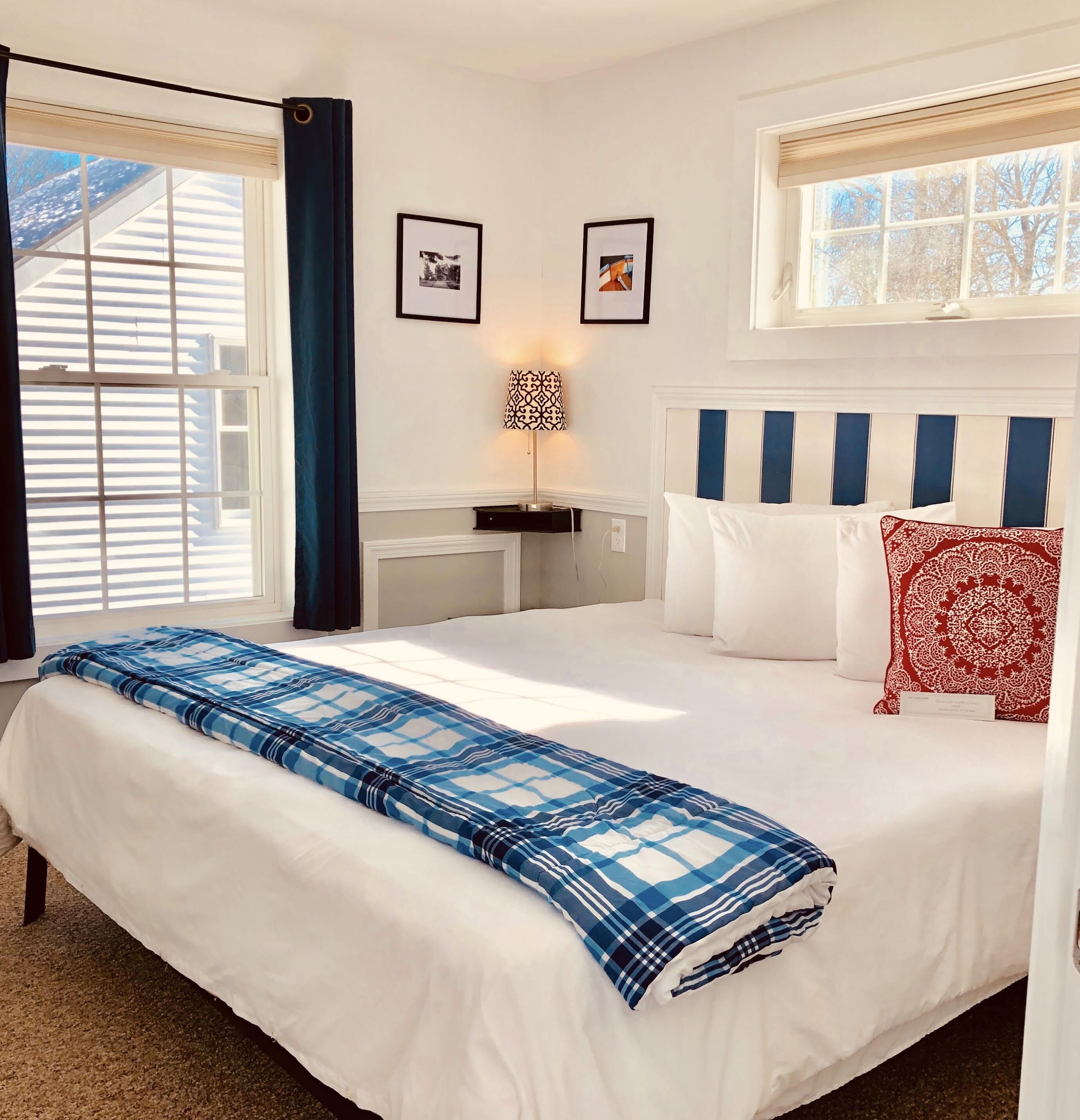 Un lit bien fait, recouvert d’une couverture à carreaux bleus et blancs, avec des oreillers blancs et un coussin à motifs rouges. Deux tableaux encadrés sont accrochés au mur, une lampe se trouve sur une table de chevet, et une fenêtre est dotée de stores.