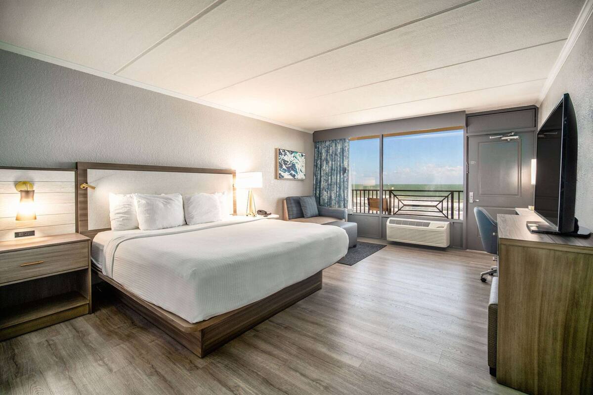 A modern hotel room with a large bed, a desk, a TV, and a balcony with a view.